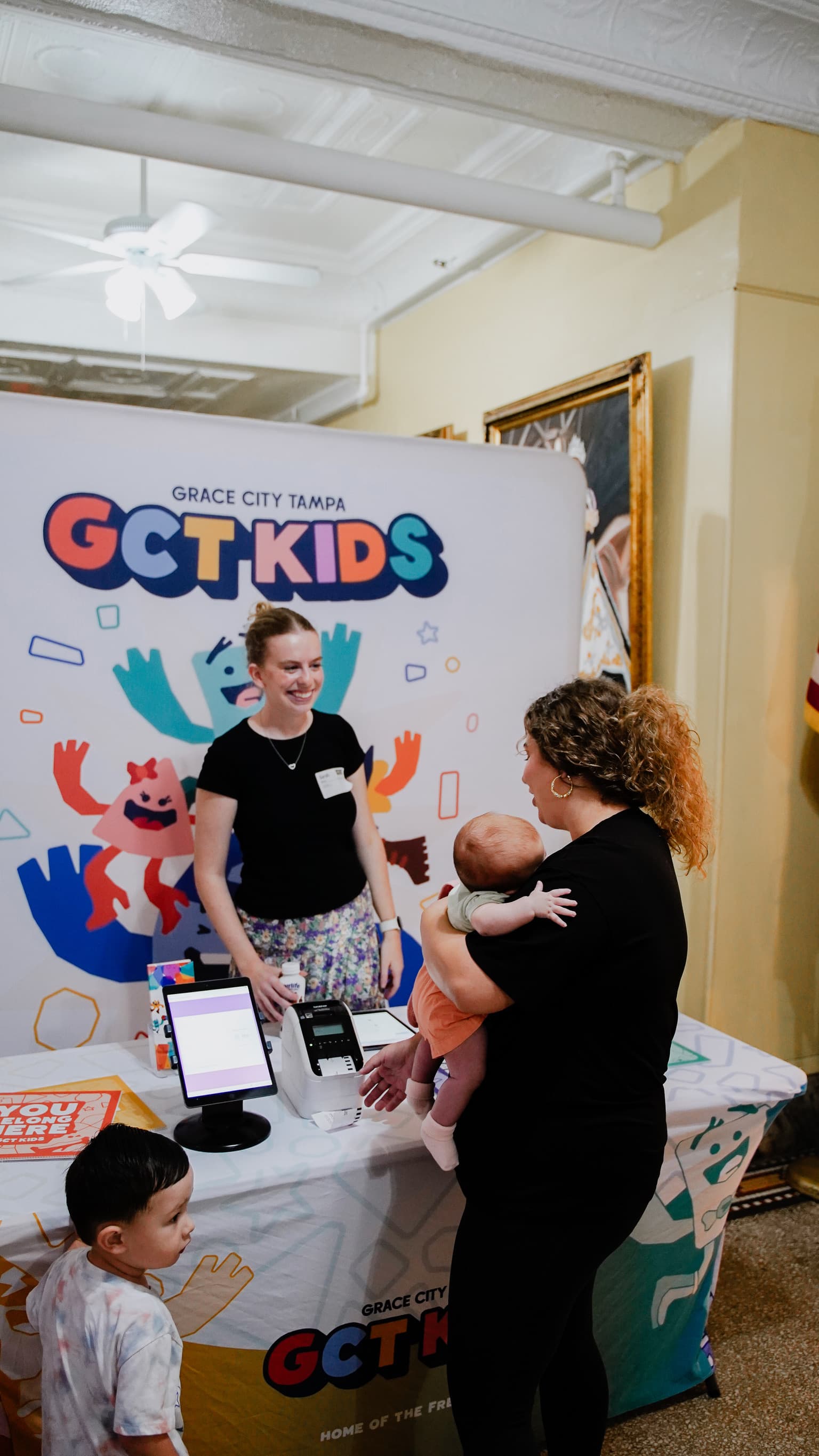 A church kids’ check-in team welcoming a family beside a printed table cover and backdrop